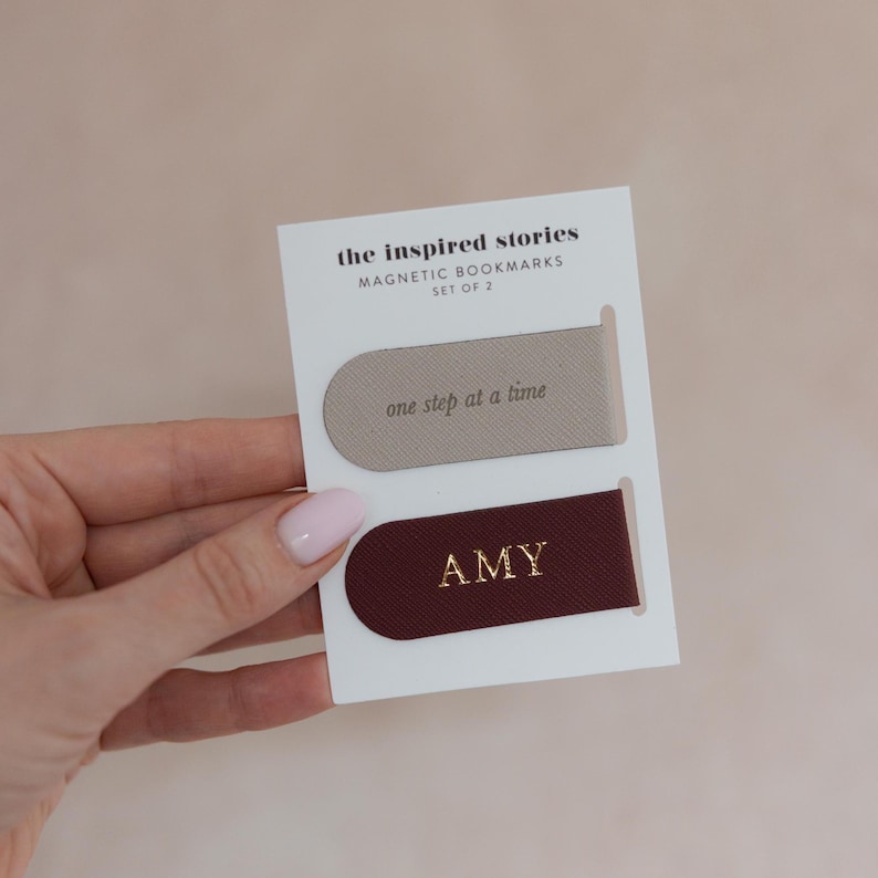 May include: A set of two magnetic bookmarks on a white card. One bookmark is taupe with the phrase "one step at a time" in gold. The other is burgundy with the name "AMY" in gold. The card reads "the inspired stories MAGNETIC BOOKMARKS SET OF 2."