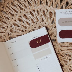 May include: A burgundy magnetic bookmark with gold initials "KL" is placed in an open planner. The planner is open to a weekly spread. A card with two magnetic bookmarks is visible, one beige and one burgundy.