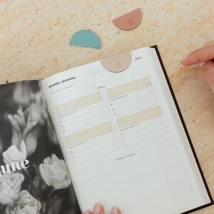 May include: Open planner with "monthly planning" and "June" text. Includes a black and white floral photo. Three half-circle shaped page markers in blue, pink, and tan are on the surface. The planner is open to a page with goal-setting sections.