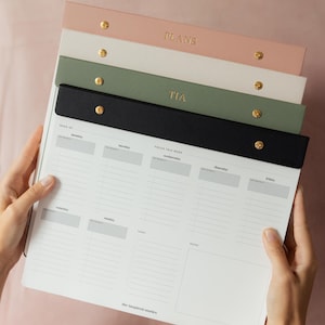 May include: A stack of weekly planners in various colors, including pink, white, green, and black. Each planner has a gold-colored fastener and the word "PLANS" or "TIA" embossed on the top. The planner pages are white with gray sections for each day of the week.
