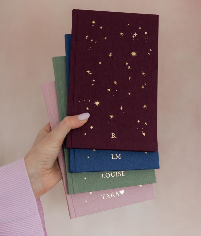 May include: A stack of personalized notebooks in various colors, held in a hand. The top notebook is burgundy with gold star constellations and the initial "B." Other notebooks are blue, green, and pink, each with a name or initials.