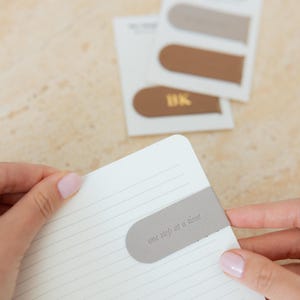 May include: A light gray bookmark with the text "one step at a time" is placed in a notebook. Other bookmarks in shades of brown and tan are visible in the background. The brown bookmark has the initials "BK" in gold.