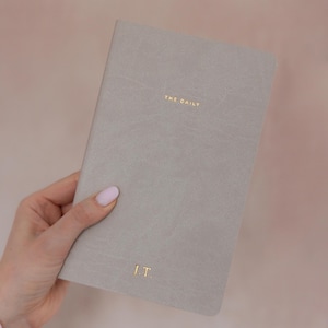 K&ouml;nnte beinhalten: Ein hellgraues Notizbuch mit einem glatten, matten Einband. Der Text THE DAILY ist in Goldfolie nahe der oberen Mitte gepr&auml;gt, und die Initialen J.T. sind in Goldfolie in der unteren Mitte auf dem Vorderdeckel des Notizbuchs eingepr&auml;gt.