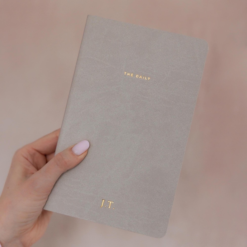 K&ouml;nnte beinhalten: Ein hellgraues Notizbuch mit einem glatten, matten Einband. Der Text THE DAILY ist in Goldfolie nahe der oberen Mitte gepr&auml;gt, und die Initialen J.T. sind in Goldfolie in der unteren Mitte auf dem Vorderdeckel des Notizbuchs eingepr&auml;gt.