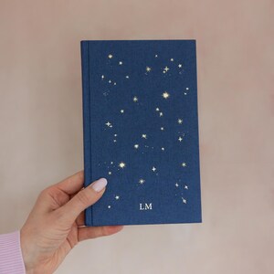 May include: A navy blue journal with a constellation design in gold foil. The cover features a scattering of stars and the initials "LM" at the bottom. The journal is held in a hand against a soft pink background.