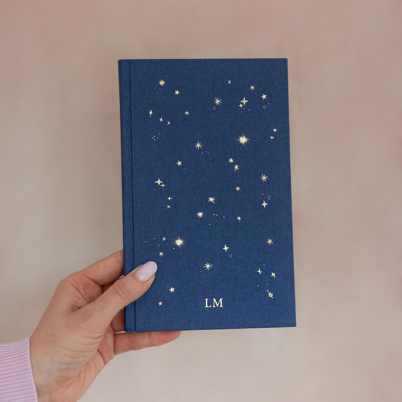 May include: A navy blue journal with a constellation design in gold foil. The cover features a scattering of stars and the initials "LM" at the bottom. The journal is held in a hand against a soft pink background.