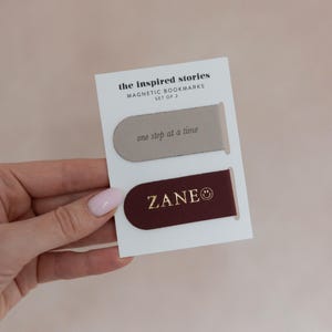 May include: A set of two magnetic bookmarks on a white card. One bookmark is taupe with "one step at a time" printed on it. The other is burgundy with "ZANE" printed on it. The card reads "the inspired stories MAGNETIC BOOKMARKS SET OF 2".
