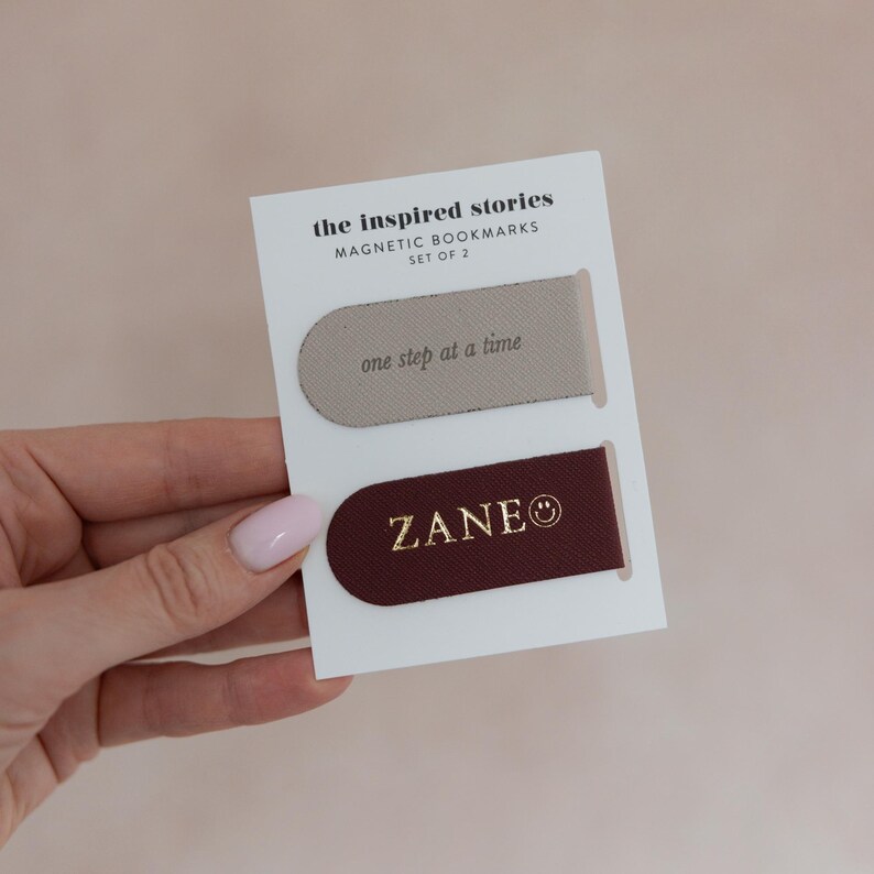 May include: A set of two magnetic bookmarks on a white card. One bookmark is taupe with "one step at a time" printed on it. The other is burgundy with "ZANE" printed on it. The card reads "the inspired stories MAGNETIC BOOKMARKS SET OF 2".