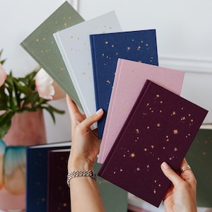 May include: A collection of hardcover journals in various colors, including green, light blue, navy, pink, and burgundy. Each journal features a gold constellation design. The journals are held in a person's hands.