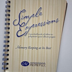 May include: A spiral-bound notebook with a light wood-grain cover. The cover features the words "Simple Expressions" in a flowing blue script, along with the text "A pocketbook of titles to jumpstart your album pages."