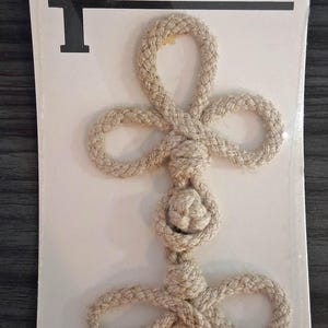 May include: A beige rope frog closure on a white display card labeled Streamline Frog Closures. The fastener features a braided cord design with two symmetrical loops at the top and bottom, connected by a central knot, all in a natural tan color.