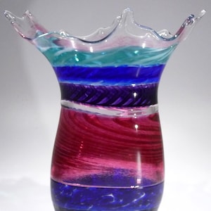 May include: A clear glass vase with a unique, splatter-like rim. The vase is decorated with colourful stripes of teal, blue, purple, and pink glass.