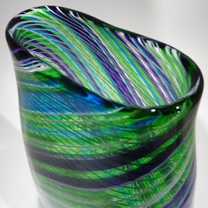 May include: A green, blue and white glass vase with a swirling pattern. The vase is made of clear glass with streaks of colour throughout.