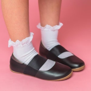May include: A pair of black leather shoes with white lace trim on the socks. The shoes have a strap across the top and a rounded toe. The shoes are worn by a person wearing a red polka dot skirt.