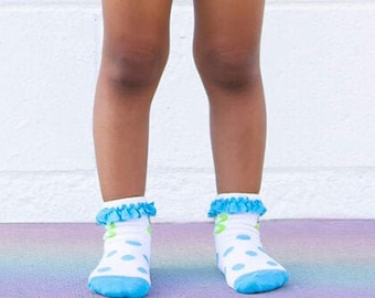 Op de afbeelding: Een jong persoon draagt een grijs T-shirt met een regenboog en het woord "hello" erop gedrukt. Ze dragen ook denim shorts, ronde zonnebrillen en blauwe en witte stippensokken.