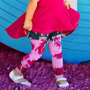 Op de afbeelding: Een jong meisje draagt een roze top zonder mouwen, een spijkerbroekje en roze tie-dye leggings. Ze staat voor een blauwe muur met witte letters die "regenboog" spellen.