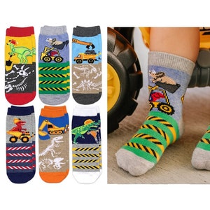 Puede incluir: Un conjunto de seis pares de calcetines con varios dise&ntilde;os coloridos de veh&iacute;culos de construcci&oacute;n y dinosaurios. Los patrones incluyen excavadoras amarillas, camiones volquete y dinosaurios verdes, junto con rayas de peligro y f&oacute;siles.