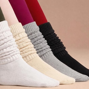 May include: A selection of slouch socks in white, cream, gray, and black. The socks are made of a soft knit material and are bunched up around the ankles. The background is a solid, neutral color.