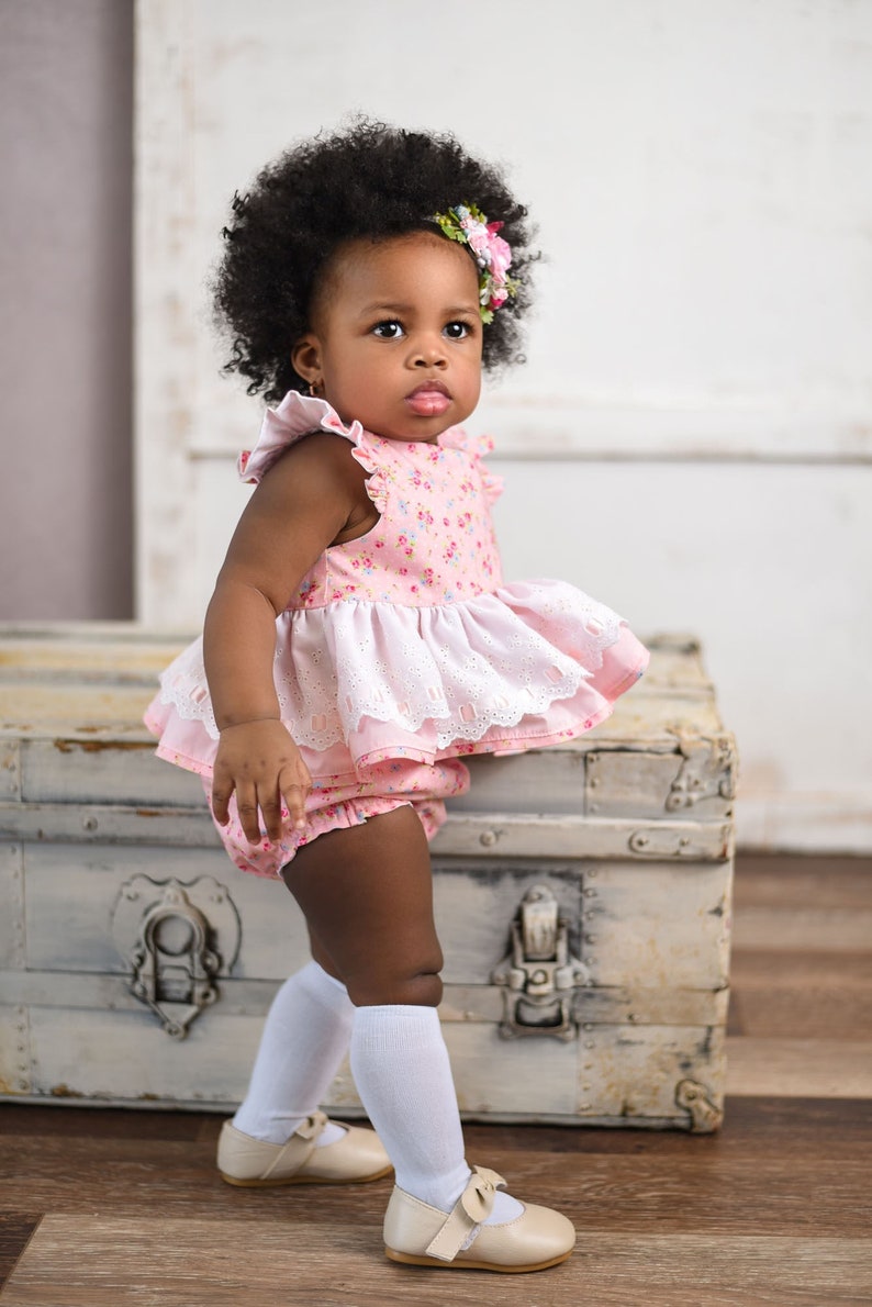Puede incluir: Una ni&ntilde;a con el pelo negro rizado lleva un vestido rosa con estampado floral y ribete de encaje blanco y calcetines blancos hasta la rodilla. Est&aacute; de pie junto a un ba&uacute;l de madera vintage.
