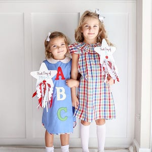Puede incluir: Dos ni&ntilde;as con vestidos sujetando carteles con forma de estrella que dicen "First Day of Pre-K" y "First Day of" con las letras A, B y C en el cartel. La ni&ntilde;a de la izquierda lleva un vestido azul con cuello blanco y calcetines blancos. La ni&ntilde;a de la derecha lleva un vestido de cuadros rojos y blancos con calcetines blancos.