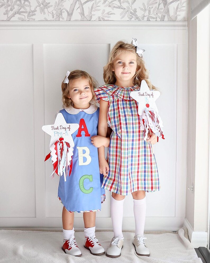 Puede incluir: Dos ni&ntilde;as con vestidos sujetando carteles con forma de estrella que dicen "First Day of Pre-K" y "First Day of" con las letras A, B y C en el cartel. La ni&ntilde;a de la izquierda lleva un vestido azul con cuello blanco y calcetines blancos. La ni&ntilde;a de la derecha lleva un vestido de cuadros rojos y blancos con calcetines blancos.