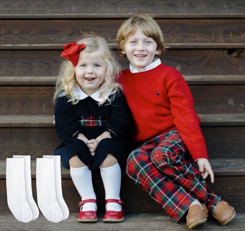 Puede incluir: Dos ni&ntilde;os, una ni&ntilde;a y un ni&ntilde;o, est&aacute;n sentados en unos escalones de madera. La ni&ntilde;a lleva un vestido negro con una faja roja y blanca a cuadros, calcetines blancos hasta la rodilla y zapatos rojos. El ni&ntilde;o lleva un jersey rojo, pantalones de cuadros y zapatos marrones. Ambos est&aacute;n sonriendo.