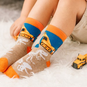 Puede incluir: Un par de calcetines de media ca&ntilde;a con base color canela, gr&aacute;ficos de esqueletos de dinosaurios en blanco, secci&oacute;n superior azul con veh&iacute;culos de construcci&oacute;n amarillos y pu&ntilde;os, talones y punteras en color naranja brillante.