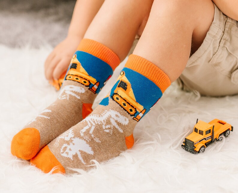 Puede incluir: Un par de calcetines de media ca&ntilde;a con base color canela, gr&aacute;ficos de esqueletos de dinosaurios en blanco, secci&oacute;n superior azul con veh&iacute;culos de construcci&oacute;n amarillos y pu&ntilde;os, talones y punteras en color naranja brillante.