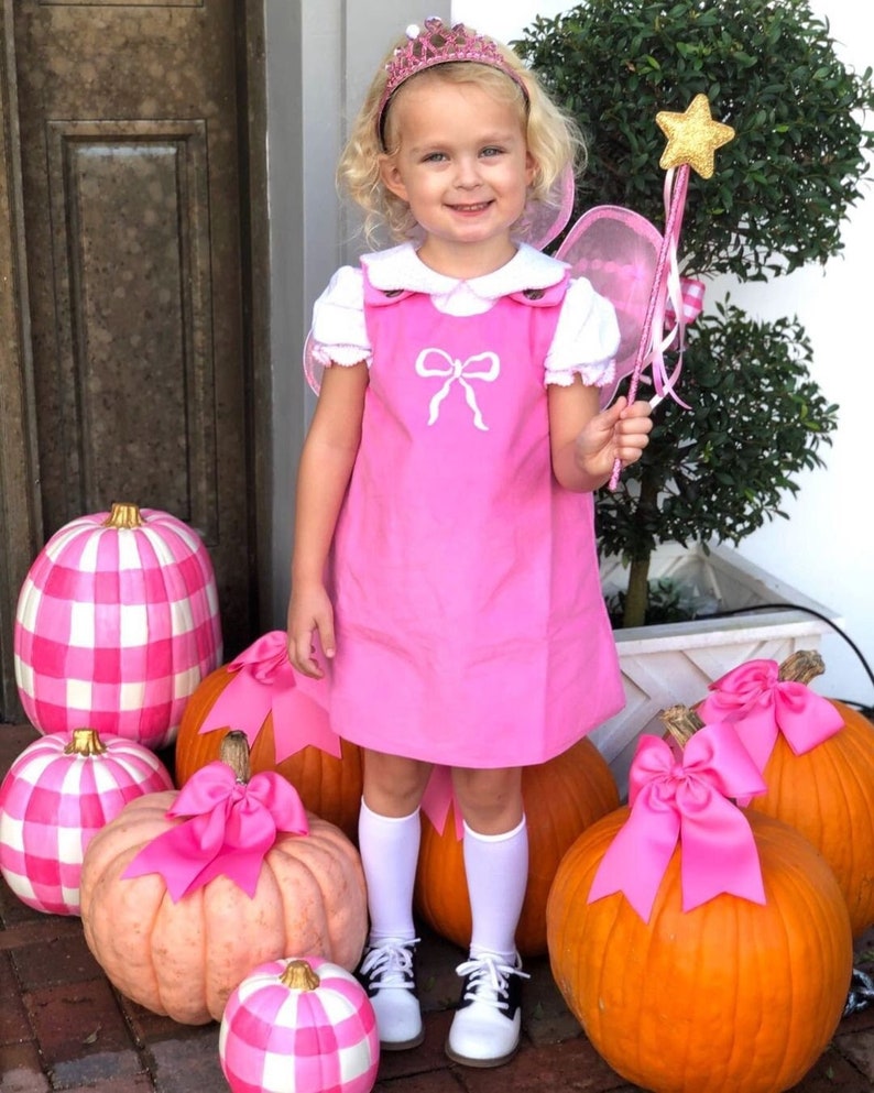 Puede incluir: Una ni&ntilde;a vestida de hada para Halloween, con un vestido rosa con cuello blanco y un lazo blanco, una tiara rosa y alas de hada. Est&aacute; sosteniendo una varita dorada en forma de estrella y est&aacute; de pie frente a una fila de calabazas decoradas con lazos rosas.