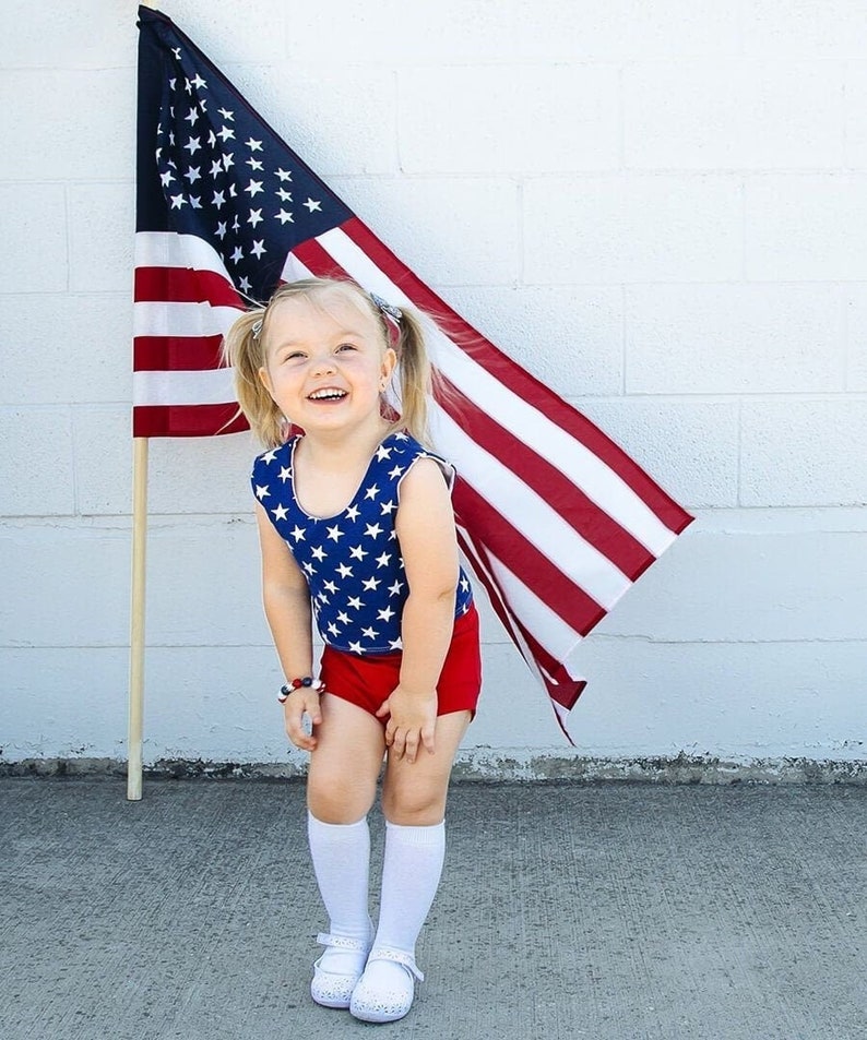 Puede incluir: Una ni&ntilde;a peque&ntilde;a con un leotardo de estrellas rojas y azules y pantalones cortos rojos se para frente a una bandera estadounidense. Ella sonr&iacute;e y lleva calcetines blancos y zapatos blancos.