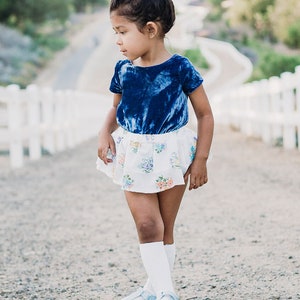 Puede incluir: Una niña pequeña con un top azul de terciopelo de manga corta, una falda blanca con estampado floral, calcetines blancos hasta la rodilla y zapatos azul claro.
