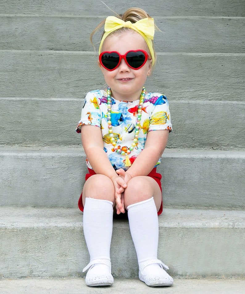 Puede incluir: Una ni&ntilde;a peque&ntilde;a con una camisa blanca de manga corta con estampado de peces de colores, gafas de sol rojas en forma de coraz&oacute;n, una diadema amarilla, pantalones cortos rojos, calcetines blancos hasta la rodilla y zapatos blancos. Est&aacute; sentada en un conjunto de escalones de hormig&oacute;n.