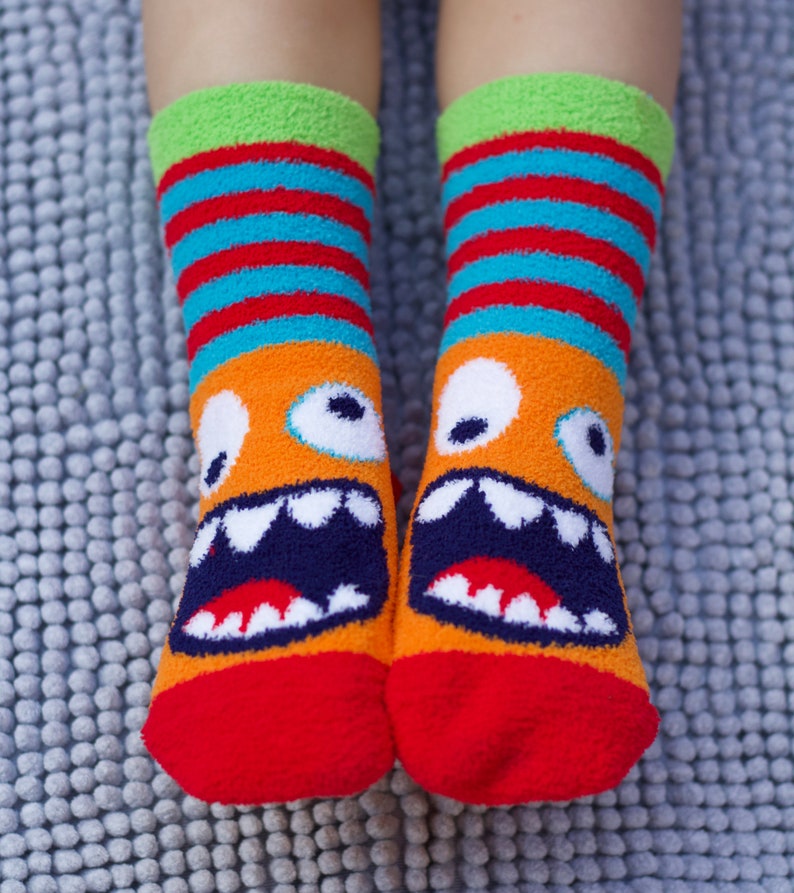 May include: A pair of red, orange, blue, and green fuzzy socks with a monster face design. The monster has white eyes, a white mouth with black teeth, and a red tongue.