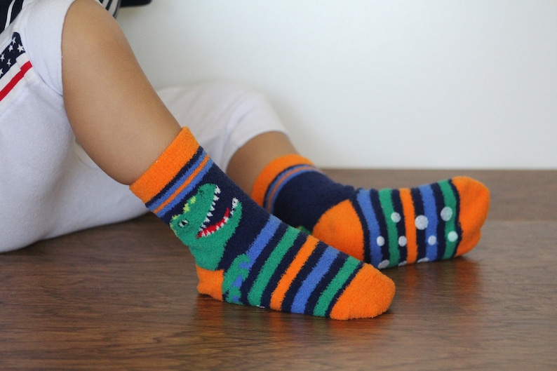 May include: A pair of colorful striped socks with a dinosaur graphic on one sock. The socks have orange, blue, green, and white stripes. One sock has white dots on the bottom.