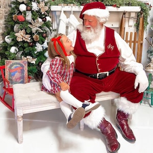 Puede incluir: Un Pap&aacute; Noel con un traje de terciopelo rojo con ribetes de piel blanca se sienta en un banco con una ni&ntilde;a peque&ntilde;a que lleva un vestido de cuadros rojos y blancos. La ni&ntilde;a sonr&iacute;e y mira a la c&aacute;mara. Pap&aacute; Noel mira a la ni&ntilde;a y tiene barba blanca. El banco es blanco y tiene un coj&iacute;n marr&oacute;n claro.