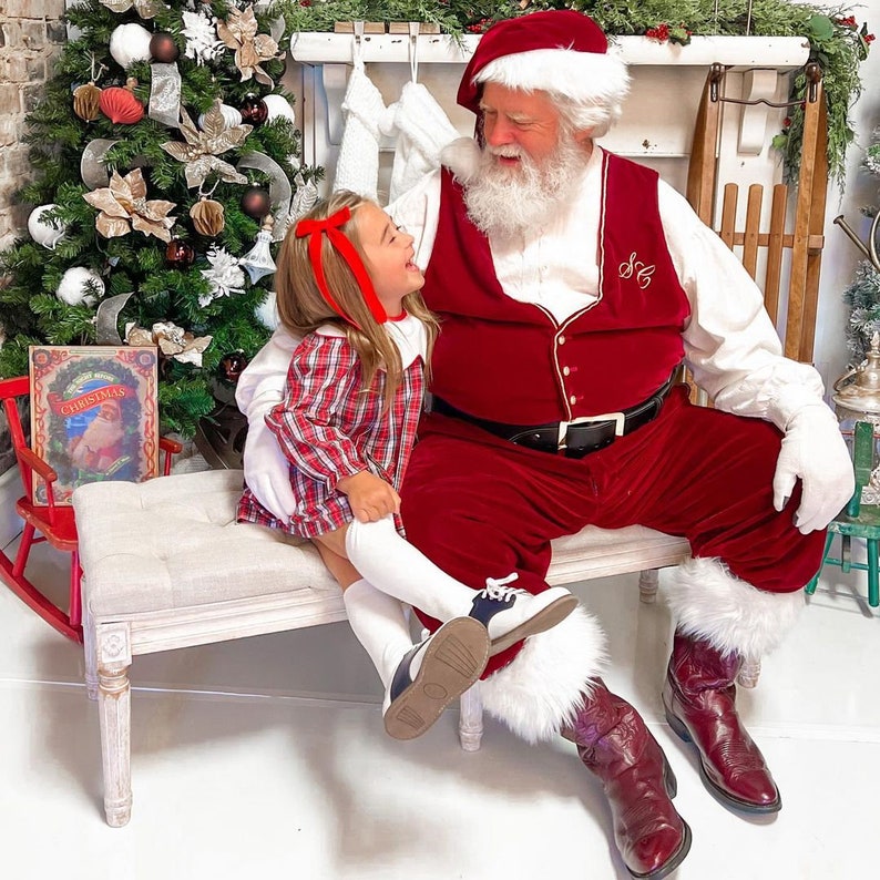 Puede incluir: Un Pap&aacute; Noel con un traje de terciopelo rojo con ribetes de piel blanca se sienta en un banco con una ni&ntilde;a peque&ntilde;a que lleva un vestido de cuadros rojos y blancos. La ni&ntilde;a sonr&iacute;e y mira a la c&aacute;mara. Pap&aacute; Noel mira a la ni&ntilde;a y tiene barba blanca. El banco es blanco y tiene un coj&iacute;n marr&oacute;n claro.