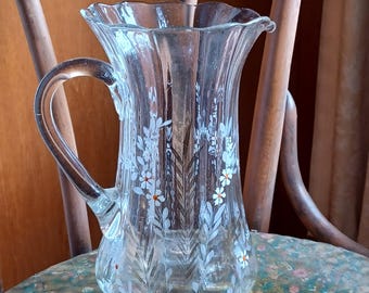 May include: A clear glass pitcher with a white floral design. The pitcher has a fluted body and a scalloped rim.