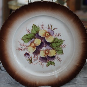 May include: A white ceramic plate with a brown rim and a floral design. The plate features a bouquet of purple and yellow pansies with green leaves. The rim has a scalloped edge.