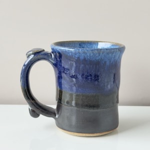 May include: A handmade ceramic mug with a dark blue and black glaze. The mug features a unique handle design and a textured surface. The mug is approximately 4 inches tall and 3 inches in diameter, perfect for coffee or tea.
