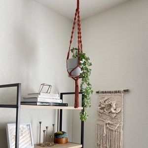 May include: A macrame plant hanger with a terracotta colored cord hangs from the ceiling with a potted plant. The plant is a trailing vine with green leaves. The hanger has a tassel at the bottom. The plant hanger is in front of a black metal shelf with books and decorative items.