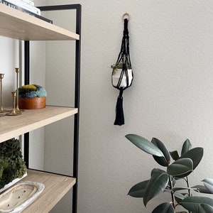 May include: A black macrame plant hanger with a wooden ring at the top, holding a white ceramic pot with a small green plant. The hanger is hanging on a white wall next to a wooden shelf with books and decor.