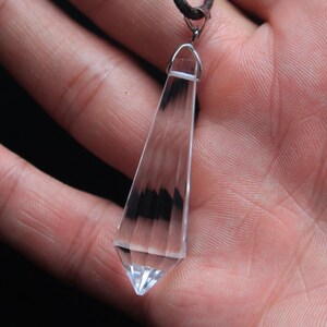 May include: A clear crystal pendant shaped like a long, pointed teardrop, hanging from a silver chain. The pendant is suspended in the palm of a hand.