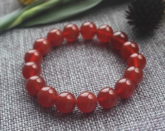 May include: A red beaded bracelet. The bracelet is made of round, polished beads in a deep red color. The beads are strung together to form a circular shape. The bracelet is displayed on a gray textured surface.