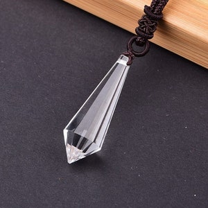 May include: A clear crystal point pendant on a brown cord necklace. The pendant is shaped like a long, thin pyramid with a pointed tip.