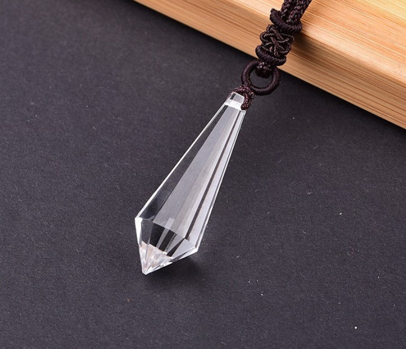 May include: A clear crystal point pendant on a brown cord necklace. The pendant is shaped like a long, thin pyramid with a pointed tip.