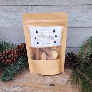 May include: A brown paper bag with a clear window showing dried apple slices, cinnamon sticks, and other spices. The bag has a label that reads "Simmer Pot Apple Pie" and instructions for use.