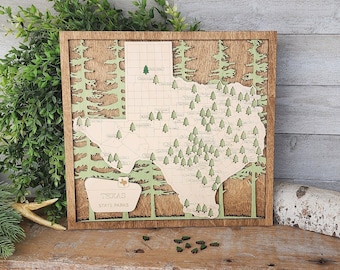 May include: A wooden map of Texas with green trees and a white background. The map is framed in a brown wood frame and is hanging on a wall. The map is a great way to show your Texas pride.