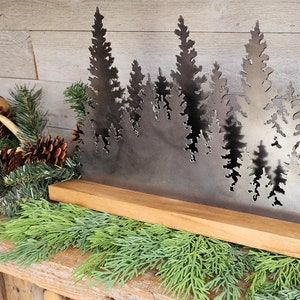 May include: A metal silhouette of a forest with multiple pine trees cut out against a silver background. The silhouette is mounted on a wooden base.