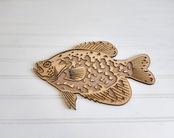 May include: A wooden cutout of a crappie fish with a detailed design. The fish is brown and has a speckled pattern.