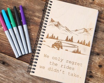 May include: A wooden spiral-bound notebook with a laser-etched mountain and off-road vehicle design. The notebook features the quote, "We only regret the rides we didn't take." Five colorful markers are arranged to the left of the notebook. The notebook is on a wooden surface.
